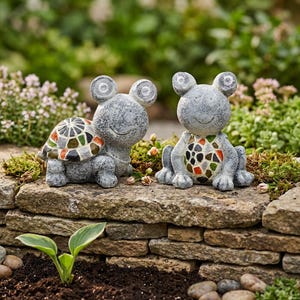 Peut inclure: Deux statues de grenouilles grises avec des motifs de coquille et de poitrine en mosaïque. Les statues ont de grands yeux ronds et sont assises sur un mur de pierre. L'arrière-plan présente des plantes et des fleurs vertes, créant une scène de jardin.