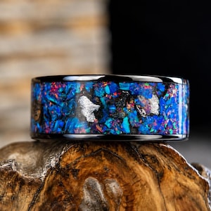 May include: A black metal ring featuring a wide inlay of crushed blue and red iridescent opal fragments interspersed with metallic silver-colored flecks. The band has a polished finish with smooth, dark edges framing the vibrant, textured inlay.