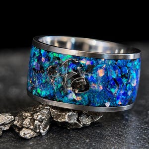 May include: A wide metal ring featuring a central inlay of crushed blue and purple iridescent stone fragments. The band has a smooth, polished silver-toned metal edge on both the top and bottom, framing the textured, colorful stone inlay.