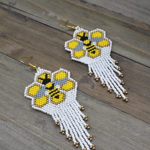 May include: A pair of white beaded earrings with a yellow and black bee design on a honeycomb background. The earrings have a long fringe of white beads.