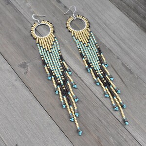 May include: A pair of long, beaded earrings with a silver-tone hoop and fish hook. The earrings feature a pattern of gold, black, and turquoise beads, creating a fringed effect. The earrings are displayed on a wooden surface.