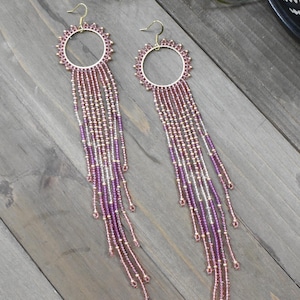 May include: A pair of long, beaded earrings. Each earring features a gold-colored hoop with a sunburst design and cascading strands of purple, gold, and clear beads. The earrings are displayed on a wooden surface.