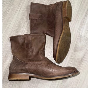 Puede incluir: Botines de cuero marrón con acabado envejecido. Las botas tienen un tacón bajo, una puntera redondeada y un detalle de correa en el lateral. Las suelas son de color marrón claro.