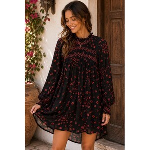 Puede incluir: Un vestido corto negro con estampado floral rojo, mangas largas con puños fruncidos, cuello alto y detalle fruncido en el corpiño. El vestido tiene una silueta holgada y fluida que cae por encima de la rodilla, confeccionado en tela ligera.