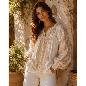 Puede incluir: Blusa color crema con bordados dorados y mangas largas abullonadas. La blusa presenta un escote en V y una botonadura frontal. El bordado dorado crea un patrón geométrico en las mangas y el corpiño. La modelo lleva pantalones blancos.