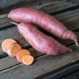 May include: Two whole elongated sweet potatoes with smooth, reddish-purple skin and four circular slices showing a bright orange interior. The raw vegetables are arranged together, displaying their natural texture and vibrant color contrast.