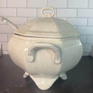 May include: A cream-colored ceramic tureen with a matching lid and a ladle. The tureen features a faceted body, two side handles, and three feet. The lid has a central handle and a beaded edge detail that matches the rim of the tureen bowl.