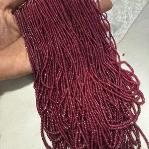 May include: A long strand of small, faceted, deep magenta-coloured gemstone beads. The beads are uniform in size and shape, featuring a reflective, sparkling surface texture that catches the light, arranged in a dense, multi-layered cluster.