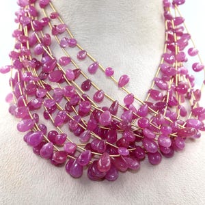 Puede incluir: Un collar de múltiples hilos con numerosas cuentas de piedras preciosas de color rosa vibrante en forma de lágrima, ensartadas en un hilo fino de tono dorado. Las cuentas están dispuestas en varias capas, creando un aspecto denso y texturizado.