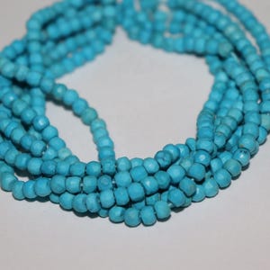 May include: A multi-strand necklace composed of numerous small, round, faceted turquoise-colored beads. The beads feature a matte finish and subtle natural veining, arranged in a circular, coiled formation with multiple overlapping strands.