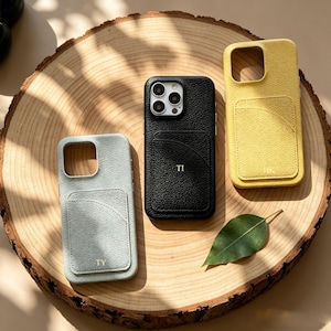 May include: Three leather phone cases in light blue, black, and yellow, each with a cardholder pocket. The black case has the initials "TI", the light blue case has "TY", and the yellow case has "JK". The cases are displayed on a wooden surface.