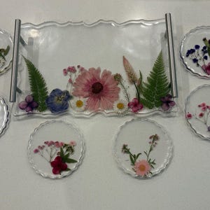 Pode incluir: Uma bandeja de servir retangular de resina transparente com alças de metal prateado e seis porta-copos circulares de resina a condizer. Cada peça apresenta flores secas embutidas, incluindo margaridas cor-de-rosa, fetos verdes e flores azuis.