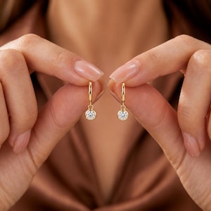 May include: A pair of gold hoop earrings, each featuring a single round, clear, bezel-set gemstone dangling from the bottom. The earrings are crafted from a polished yellow metal, showcasing a simple, refined design with a secure circular hoop.