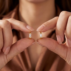 May include: A pair of small gold hoop earrings, each featuring a single clear baguette-cut gemstone set vertically in the center. The earrings have a polished metallic finish and a hinged closure mechanism, showcasing a simple and refined design.