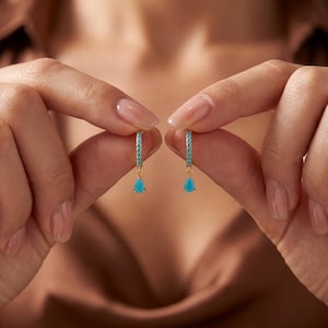 May include: A pair of gold hoop earrings featuring a row of small, bright turquoise stones along the hoop and a single, dangling, pear-shaped turquoise drop charm suspended from the base of each earring, creating a delicate and refined appearance.