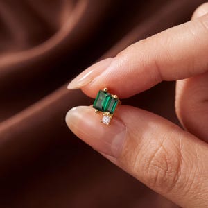 May include: A gold stud earring featuring two rectangular emerald green stones arranged in a staggered formation, accented by a single small round clear stone at the base, all set in a polished gold metal frame with a secure post backing.