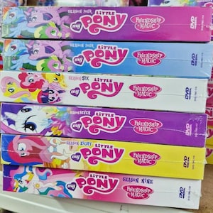 Könnte beinhalten: Ein Stapel DVD-Boxen der Zeichentrickserie "My Little Pony: Freundschaft ist Magie". Die Boxen sind in verschiedenen Farben gehalten und zeigen das Logo der Serie sowie die Staffeln. Auf jeder DVD-Hülle steht "Friendship is Magic".
