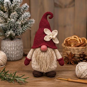 May include: A crocheted gnome figurine featuring a tall, pointed dark red hat adorned with a cream-colored flower and berries. The gnome has a round pink nose, a long beige yarn beard, a cream-colored textured body, dark red mittens, and brown feet.