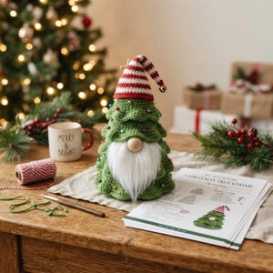 May include: A crocheted Christmas tree gnome with a green tiered body, a red and white striped hat, a white faux fur beard, and a wooden nose. The gnome is decorated with small colorful beads and sits next to a printed crochet pattern document.