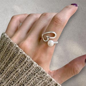 Könnte beinhalten: Ein silberfarbener Ring mit einem offenen, herzförmigen Drahtdesign auf der einen Seite und einer einzelnen, glatten, runden weißen Perle auf der anderen Seite. Die Ringschiene besteht aus poliertem Metall, das sich zum offenen Herz formt.