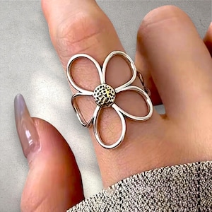 Könnte beinhalten: Ein silberfarbener Ring mit einem großen, offen gestalteten Blumenmotiv. Die fünf Blütenblätter bestehen aus dünnen, glatten Metallschleifen, während die Mitte der Blume eine texturierte, kreisförmige Oberfläche mit einem dunkleren Metallfinish aufweist.