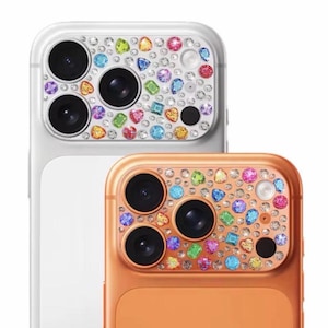Puede incluir: Dos protectores de lentes de cámara para teléfonos inteligentes. Uno es blanco, el otro naranja. Ambos están decorados con pedrería y gemas de colores. Las lentes son negras.