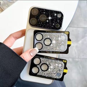 Op de afbeelding: Een smartphone met een zwarte glitter cameralensbeschermer geïnstalleerd, vastgehouden naast twee extra zilveren glitter cameralensbeschermers met het label IP 17 PRO MAX en 9H. De beschermers hebben ronde uitsparingen voor de lenzen.