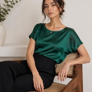 May include: A dark emerald green short-sleeved blouse with a smooth, satin-like finish, a rounded neckline, and a relaxed fit, paired with solid black trousers. The blouse features a subtle sheen and short, loose sleeves that drape over the shoulders.