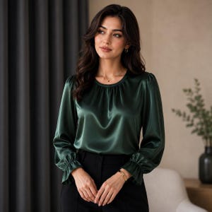 May include: A dark forest green long-sleeve blouse with a satin-like finish, featuring a round neckline with subtle gathering, and sleeves that end in ruffled cuffs. The blouse is paired with black trousers, creating a sleek and polished appearance.
