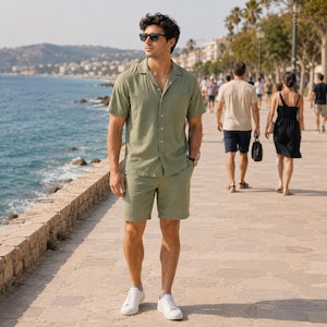 May include: A sage green short-sleeved button-down shirt with a notched collar and matching sage green shorts. The outfit is paired with white low-top sneakers and black sunglasses. The clothing features a textured fabric finish and a relaxed fit.