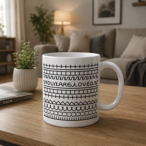 Puede incluir: Taza de cerámica blanca con un patrón geométrico negro y la frase "YOU ARE LOVED". La taza tiene un asa curvada. Está sobre una mesa de madera. La taza es de tamaño estándar.