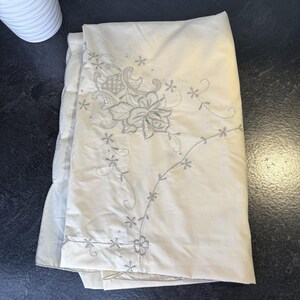 May include: Cream-colored bed sheet with embroidered floral and vine design in gray. The embroidery features a large flower and smaller star-shaped flowers. The sheet is folded, showing the detailed stitching and texture of the fabric.