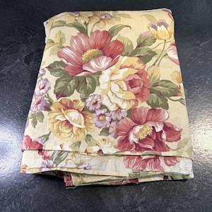May include: Folded floral fabric featuring large, colorful blooms in shades of red, yellow, and pink, set against a cream background. The fabric has a classic, vintage aesthetic, with detailed floral patterns and green leaves.