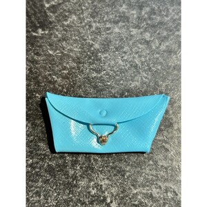 May include: A light blue, textured pouch with a curved flap and a gold-colored clasp. The pouch is closed with a small, round button. The background is a dark, speckled surface.