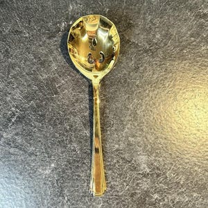 May include: A polished gold-tone serving spoon featuring a wide, rounded bowl with two distinct, symmetrical cut-out shapes near the center. The handle is slender and straight, transitioning into a flared base with vertical grooved detailing.