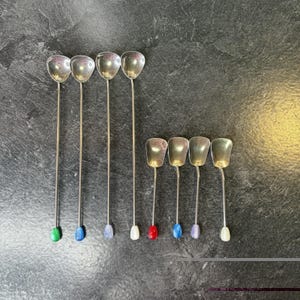 May include: A set of eight metal spoons with long, slender handles. Four spoons are longer with green, blue, light blue, and white tips. Four shorter spoons feature red, blue, light blue, and white tips. The bowls are metallic silver.