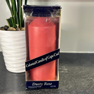 May include: A Dusty Rose unscented pillar candle in a clear plastic box. The candle is a solid, cylindrical shape with a smooth surface. The box has a black label that reads "Colonial Candle of Cape Cod" and "Dusty Rose."
