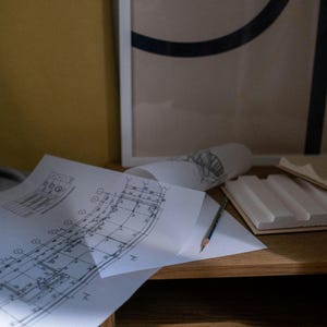 Op de afbeelding: Gedetailleerde architectuurtekeningen en opgerolde tekeningen liggen op een houten bureau, samen met een potlood en een wit schuimmodel. Een abstract kunstwerk in een lijst hangt op de achtergrond.