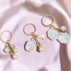May include: Three gold keychains with pearl and shell-shaped charms. Each keychain has a gold ring, a small tag, and a cluster of pearls or shell-shaped pendants. The keychains are arranged on a pink satin surface.