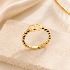 May include: A gold-toned ring featuring a beaded band design and a central emblem with the letters CD. The ring has a polished metallic finish and a circular shape, showcasing a consistent bead pattern around the entire circumference of the band.