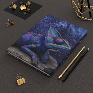 May include: A hardcover notebook featuring a vibrant, multi-colored chameleon resting on a rock against a dark, starry night sky background. The notebook is accompanied by a gold-colored pen, a black pen, and several gold-toned metal binder clips.