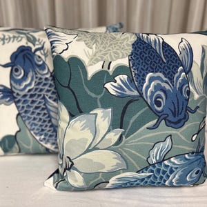 May include: Two square throw pillows with a blue and white koi fish design. The pillows feature a detailed pattern of koi fish swimming among aquatic plants in shades of blue, teal, and white. The background is a light cream color.