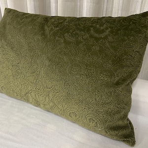 May include: Olive green rectangular pillow with a floral pattern. The pillow has a soft, velvety texture and a detailed embossed design. The pillow is placed on a white surface.