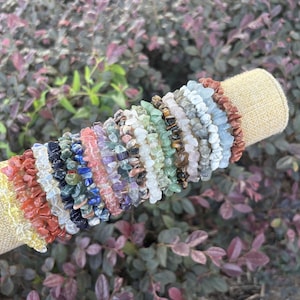 May include: A collection of multiple stretch bracelets made from small, irregular tumbled gemstone chips in a variety of colors including red, blue, green, yellow, clear, and brown, displayed on a cylindrical beige fabric jewelry holder.