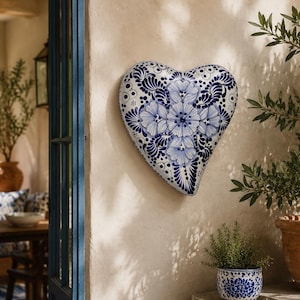 Puede incluir: Un adorno de pared de cerámica en forma de corazón con un diseño floral azul y blanco. La pieza decorativa está montada en una pared de color claro, con una vista de un patio exterior en el fondo. El corazón mide unos 30 cm de ancho.
