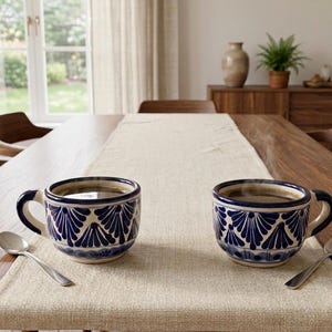 Puede incluir: Dos tazas de cerámica azul y blanca llenas de café están sobre una mesa de madera, cada una con una cuchara de plata. Las tazas tienen un patrón decorativo. También se ve un camino de mesa beige y sillas de madera.