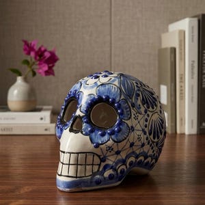 Puede incluir: Calavera de cerámica con un diseño floral azul y blanco. La calavera tiene cuencas de ojos grandes y una boca sonriente detallada. La pieza decorativa está colocada sobre una superficie de madera, con libros y un jarrón de flores en el fondo.