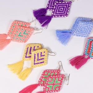Puede incluir: Una colección de pendientes coloridos hechos a mano con diseños cuadrados tejidos y borlas a juego. Los pendientes presentan varias combinaciones de colores, incluyendo rosa, morado, azul, amarillo y verde, con ganchos para las orejas plateados.