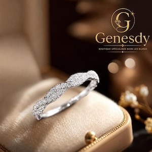 Peut inclure: Une bague argentée présentant un design de bande ondulée et torsadée incrustée de nombreuses petites pierres claires et scintillantes. La bague est placée dans une boîte à bijoux doublée de velours beige. Le texte Genesdy est visible en haut à droite.