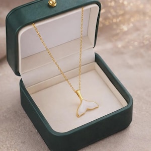 Può includere: Una collana color oro con un ciondolo a forma di coda di balena bianco e oro, presentata in un portagioie verde scuro aperto con interno bianco. La catena della collana è delicata e il ciondolo è dettagliato.